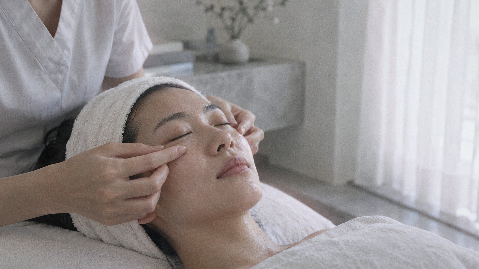 Therapist's fingertips pressing gently along the under-eye area of a woman lying on a facial bed